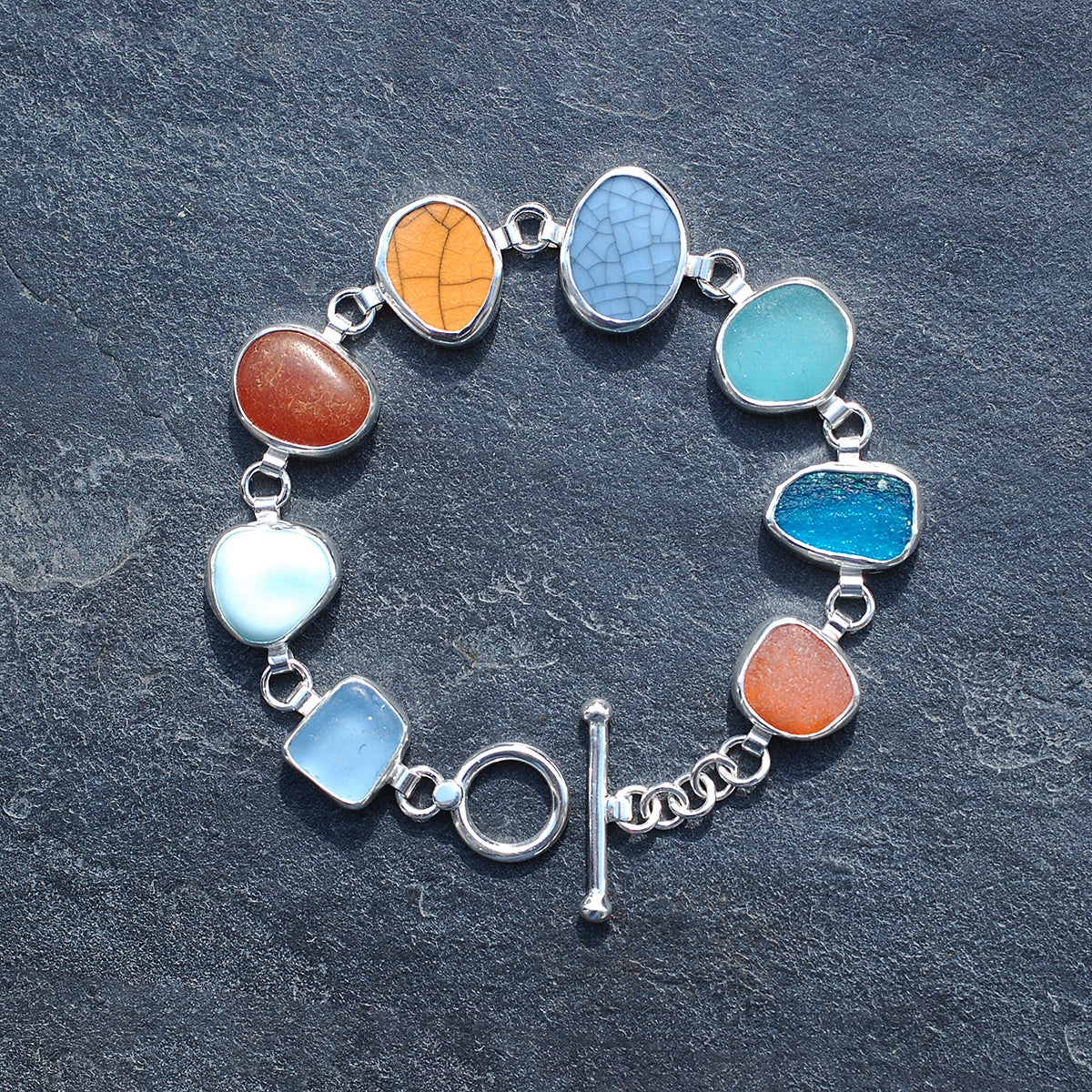 Silver bezel set bracelet with sea glass and pottery pieces on a grey slate background.