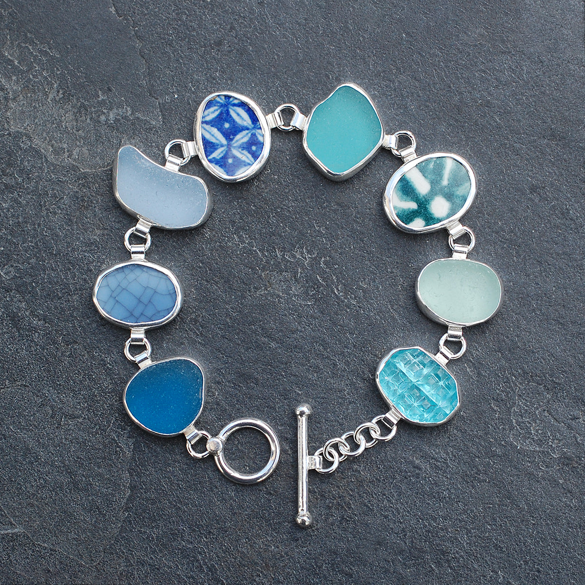 Blue and green sea glass and pottery shard bracelet on a slate grey background.