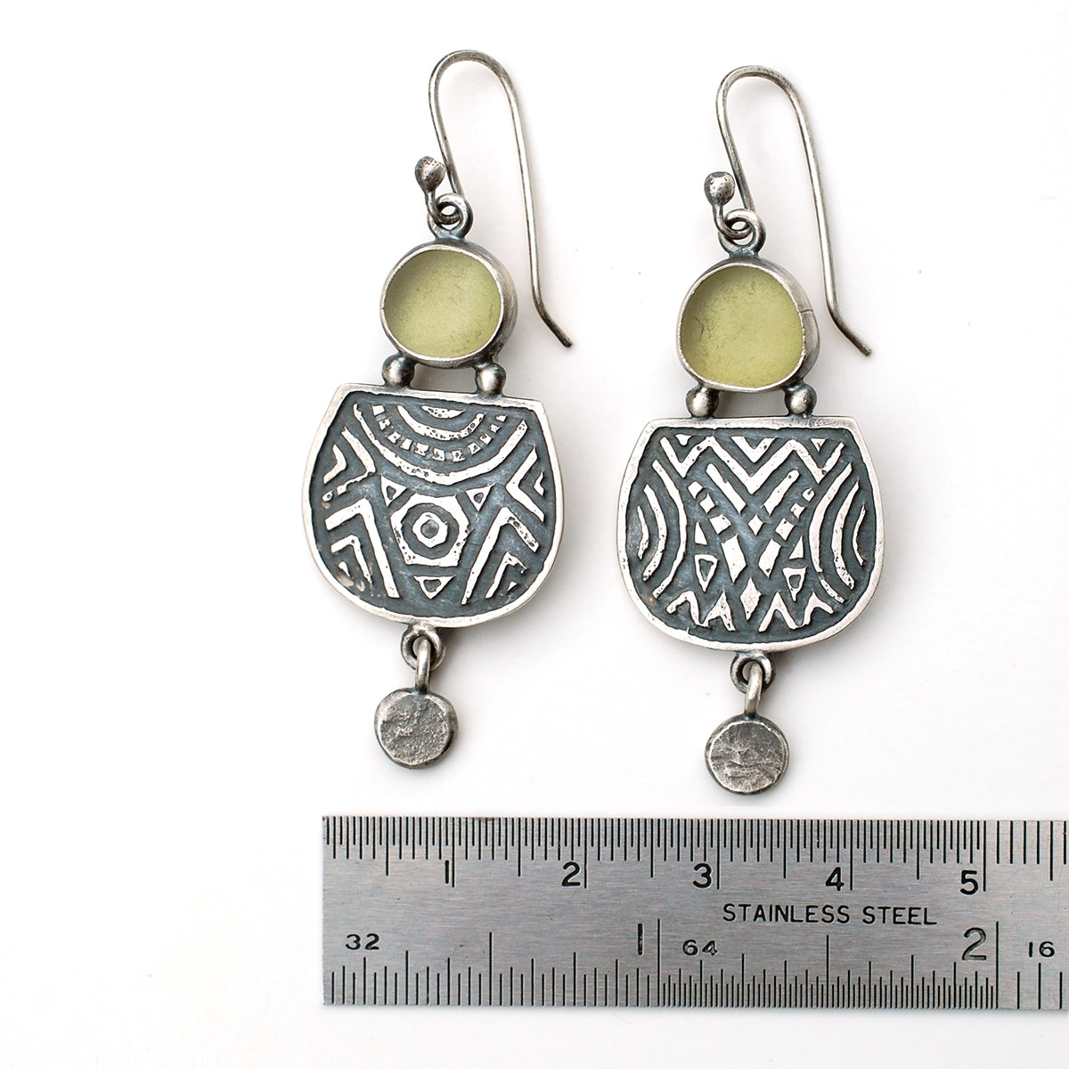 Etched silver earrings with olive green sea glass,  on a white background, with a ruler